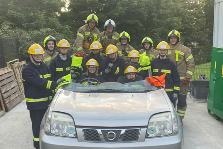 Pupils in fire fighter equipment gather around a car cut in half