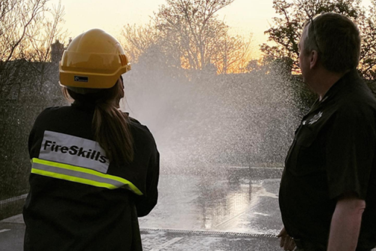 A pupil in fire fighter equipment using a fire hose beside an instructor